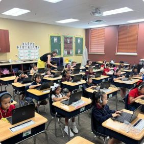 Elementary students working on laptops in a classroom with a teacher assisting