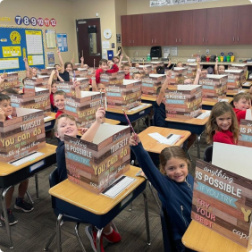 Students in a classroom holding up project boxes and smiling at their desks